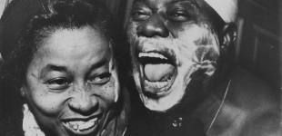 Louis Armstrong with cloth around head and shaving cream on face with Lil Hardin Armstrong; Lil was visiting Louis backstage; (another shot from this set was used in EBONY)