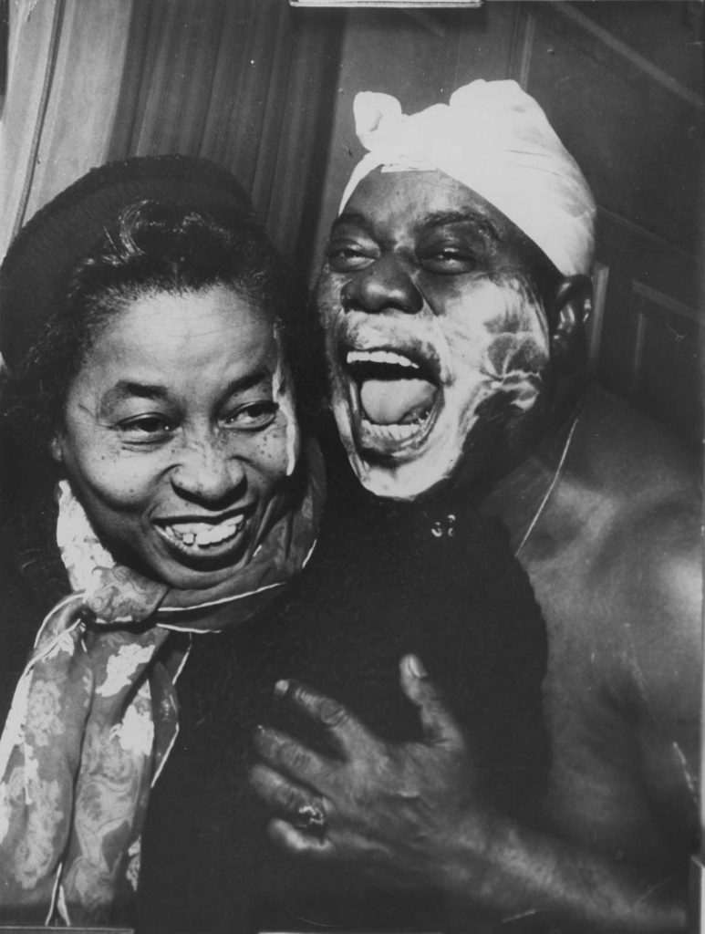 Louis Armstrong with cloth around head and shaving cream on face with Lil Hardin Armstrong; Lil was visiting Louis backstage; (another shot from this set was used in EBONY)